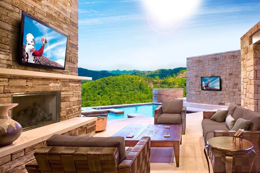 A backyard with a sitting area and two Séura outdoor TVs, one above a fireplace and another on a wall by the pool.