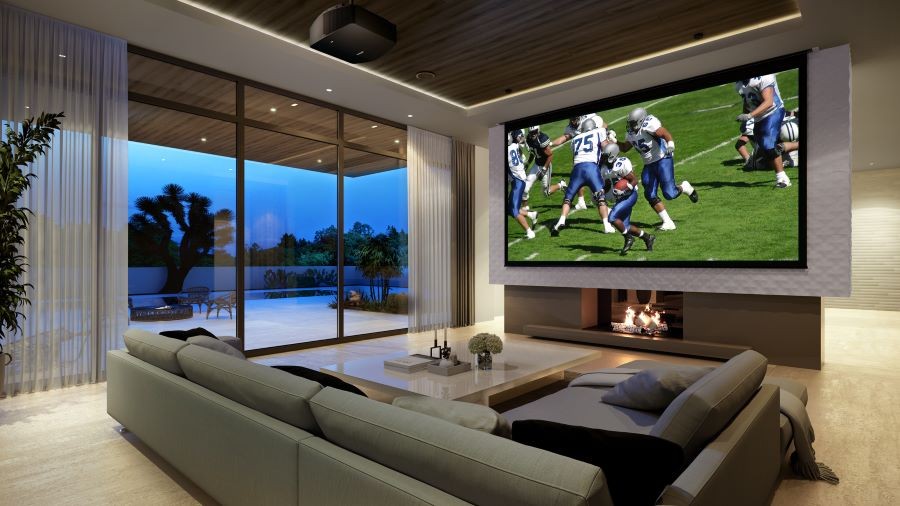 A living area with a sectional, Sony projector, and a large screen showing a football game above a fireplace.