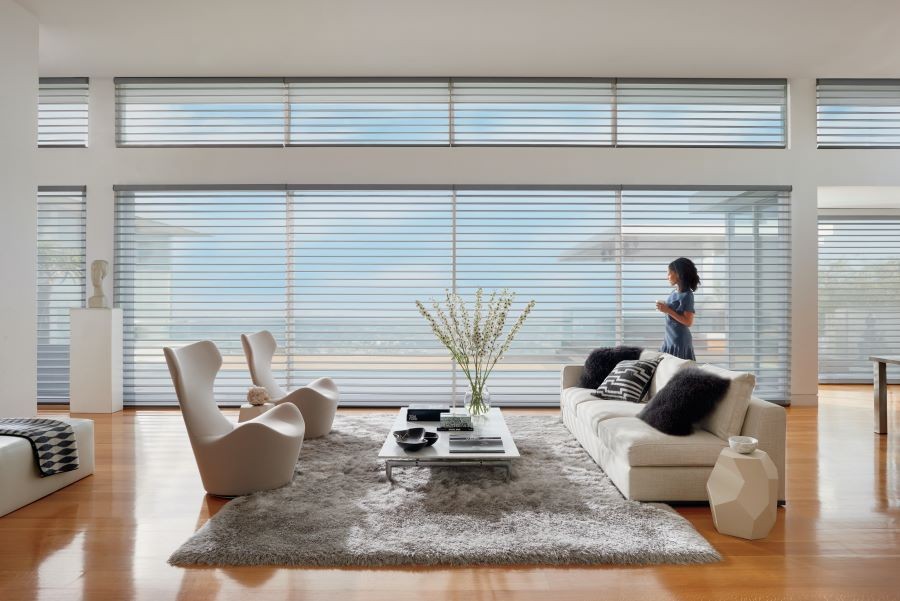 A woman in a living room looking out picture windows covered by Hunter Douglas motorized blinds.