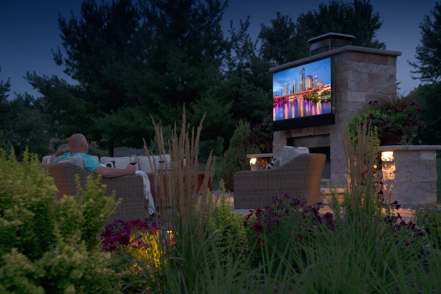 Two people are watching a Seura outdoor TV at night in a garden area.