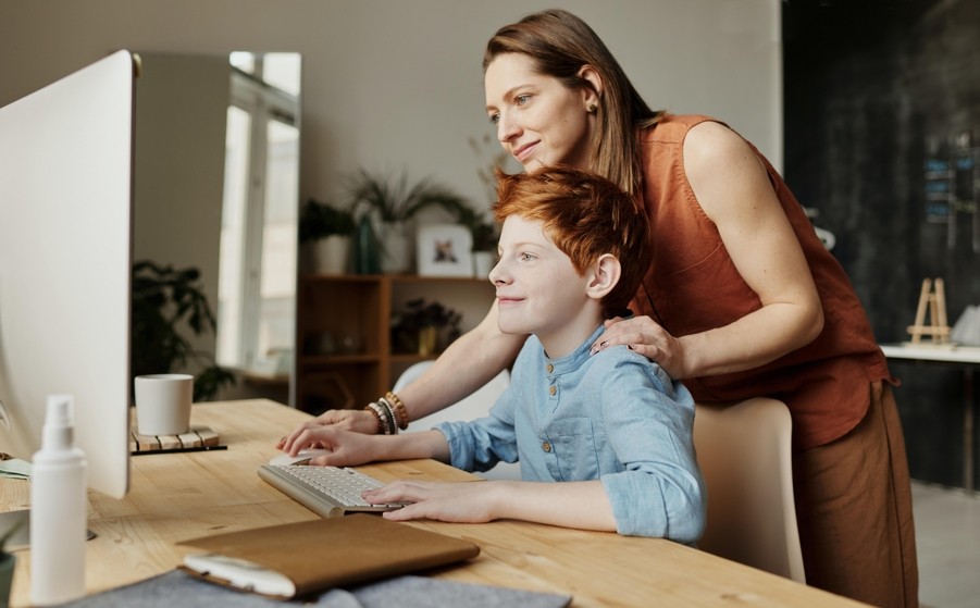 A smiling woman helps her son do homework on a desktop computer at home.