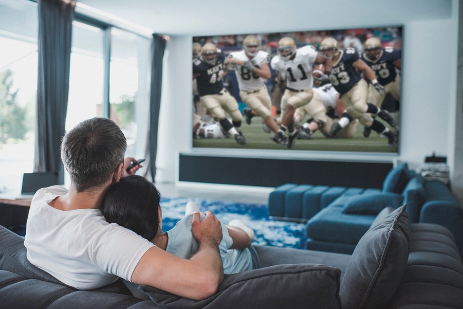 Two people sit on a sectional sofa watching a football game on a wall-mounted TV display.